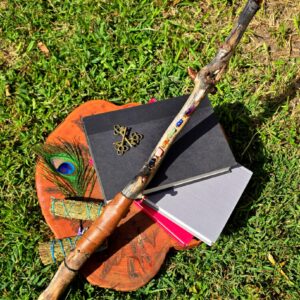 A handcrafted ritual wand made of carved wood, adorned with crystals, feathers, and bones. The background of the photo is a slab of wood with books and notebooks laying across it, a bundle of sage to the left, and the wands laying across the top.